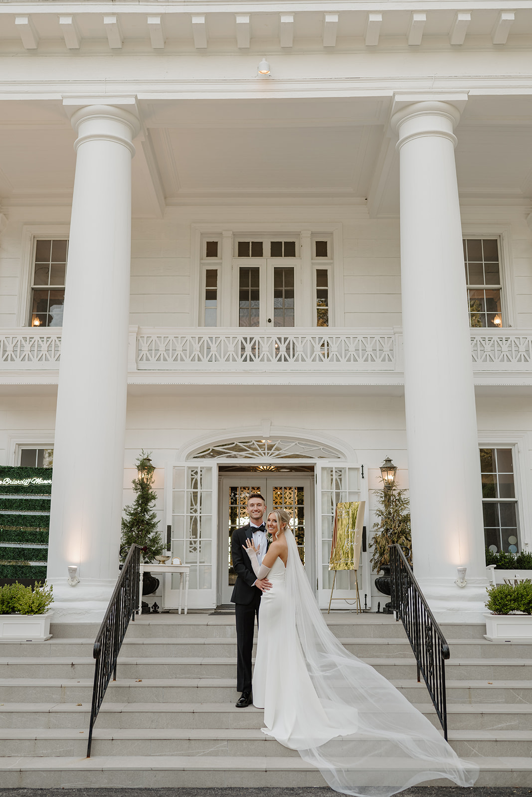 Bride and groom pose on mansion steps, with the bride's long, sheer veil flowing down the stone stairs.